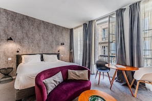 Een moderne hotelkamer met een groot bed, een paarse bank, een ronde houten tafel en uitzicht op een gebouw door dunne gordijnen.