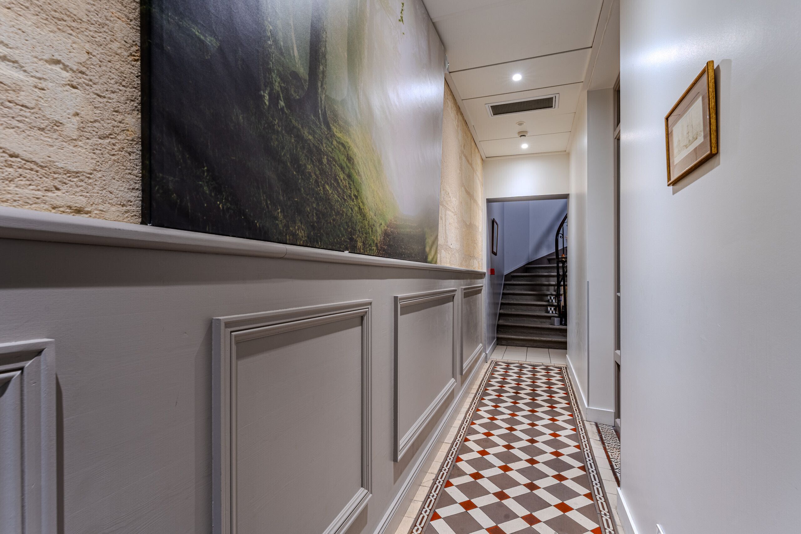 Un pasillo con piso a cuadros, un cuadro grande en la pared y una escalera al fondo.