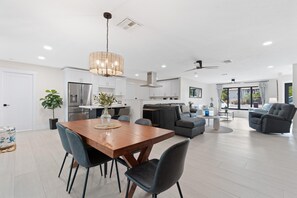 A modern kitchen with white cabinets, a dining area with a wooden table and chairs, a living room with a sofa and a coffee table, and a large window with curtains. - Sunshine Dock House (Fort Myers Beach)