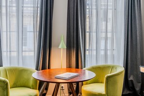 Ein Zimmer mit zwei grünen Sesseln, einem Holztisch, einer grünen Stehlampe und einem Fenster mit durchsichtigen Vorhängen.