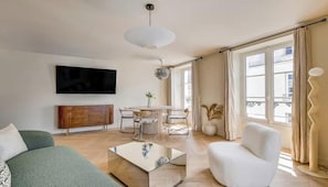 Una sala moderna con un televisor de pantalla plana, una mesa de comedor con sillas, un sofá, un sillón blanco y un gran ventanal con cortinas.