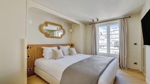 Une chambre à coucher avec un lit, un miroir, une fenêtre avec des rideaux et une vue sur des immeubles.