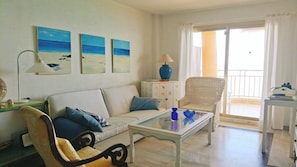 A living room with a sofa, armchair, glass coffee table, and a wicker chair. There are three paintings on the wall, a lamp, and a cabinet with a starfish design. - Apartment in front of the sea with beautiful views. The beach just below (Fuengirola)