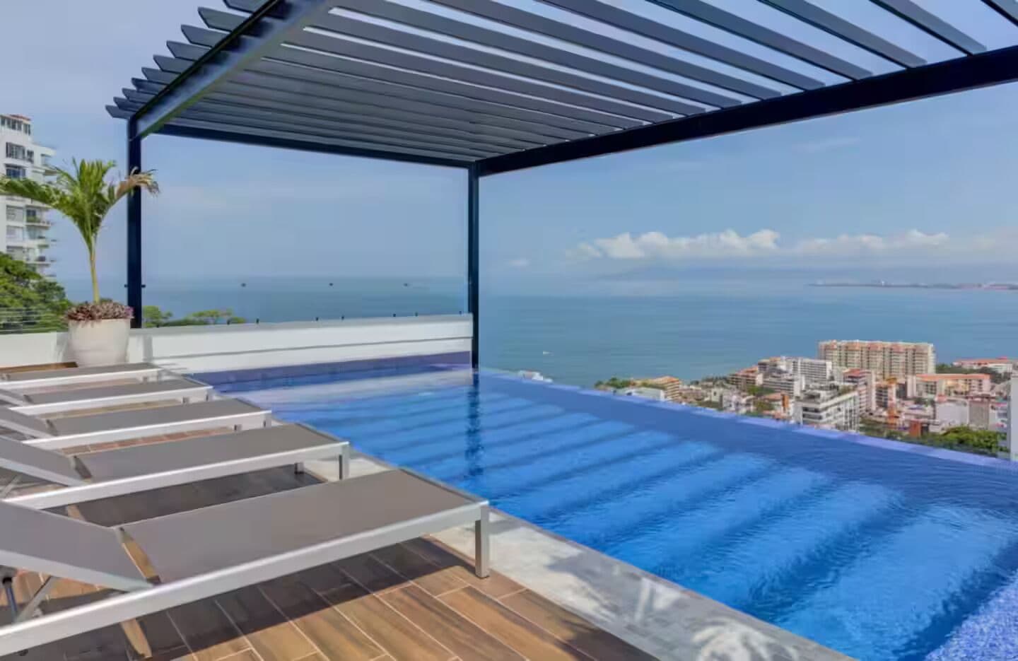 A rooftop infinity pool with lounge chairs, a pergola, and a cityscape view.