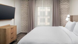 A hotel room with a bed, a wooden dresser, a TV mounted on the wall, and a brick-patterned wall. - Accessible 1BR Suite | Hot Breakfast & Pool Access (San Antonio)