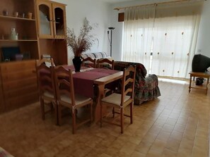 Een eetgedeelte met een houten tafel en stoelen, een kast met planken, een potplant en een bank.