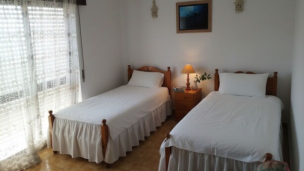 Quarto com duas camas de solteiro, mesa de cabeceira com abajur, janela com cortinas de renda e um quadro na parede.