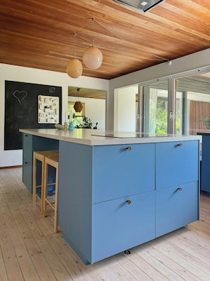 Eine moderne Küche mit einer blauen Kochinsel, Holzboden und Pendelleuchten, mit Blick durch große Fenster.