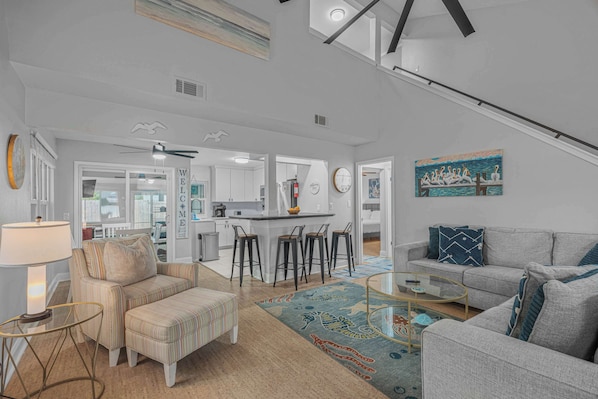 A spacious living area with a sofa, ottoman, and a coffee table. There is a kitchen in the background with a bar stool and a painting on the wall. - 3BR Home, Indoor Pool, Hot Tub, Dog Friendly (Gulf Breeze)