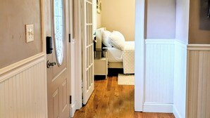 Een hal met houten vloer, een deur met ruit en een slaapkamer met bed en kledingkast.