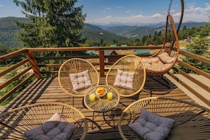 Une terrasse en bois, agrémentée de meubles en osier, dont des chaises et un fauteuil suspendu, offrant une vue sur un paysage montagneux.