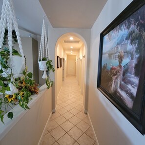 A hallway with a tiled floor, a white macrame hanging plant, a framed painting, and a door at the end. - Beautiful 4,000 sq ft home at the base of the Colorado National Monument. (Grand Junction)