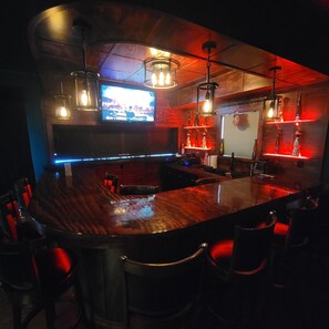 A bar area with a curved wooden bar, hanging pendant lights, and a television screen. - Beautiful 4,000 sq ft home at the base of the Colorado National Monument. (Grand Junction)