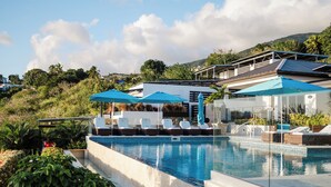 A swimming pool with lounge chairs and umbrellas, surrounded by greenery and hills.