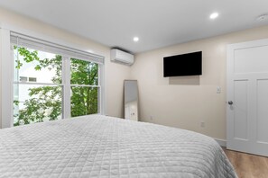 A bedroom with a large bed, a flat-screen TV mounted on the wall, a window with a view of trees, and an air conditioning unit. - 3BR near Acadia & Bar Harbor sleeps 8 - AC - WiFi - Deck (Ellsworth)