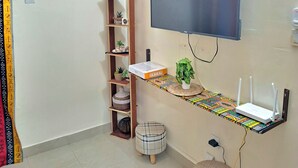 Ett vardagsrum med en platt-TV som är monterad på väggen, en trähylla med dekorativa föremål, en rund pall och ett litet bord med en router och prydnadsväxter.