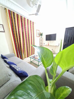 A living room with a sofa, a coffee table, a plant, curtains, a ceiling fan, a TV, and a door. - Téranga Home Cosy, 600 meters from the beach (DAKAR)