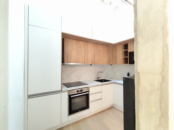 Une cuisine compacte avec des meubles blancs, un four encastré, un évier et des placards en bois situés en hauteur.