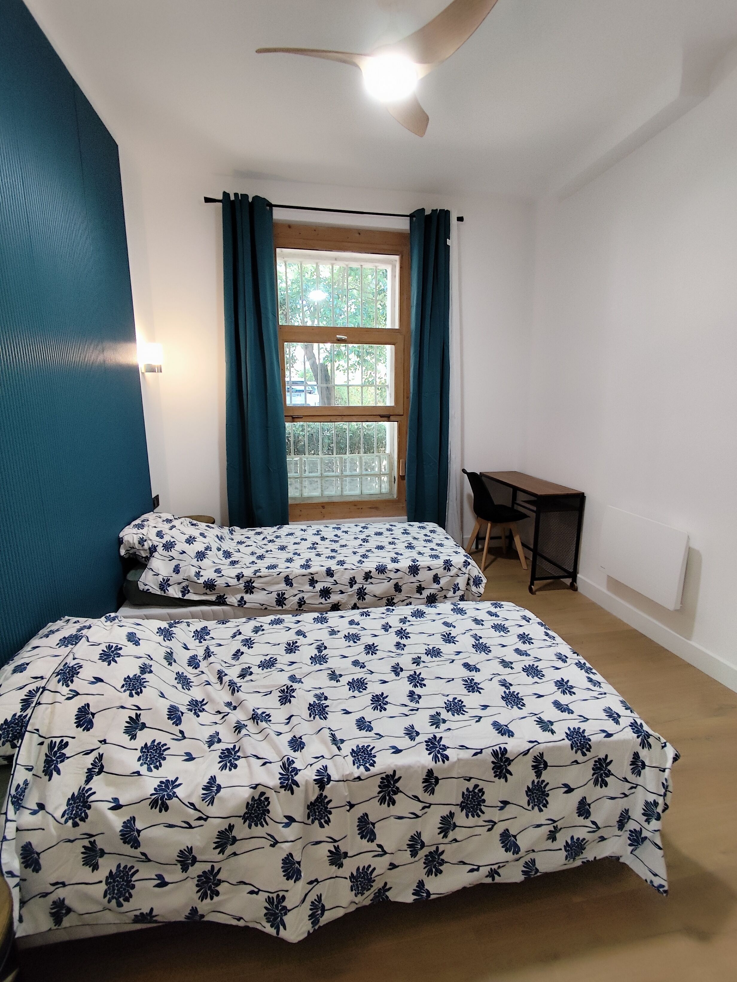 A bedroom with a bed, a desk, a chair, a window with curtains, and a ceiling fan.