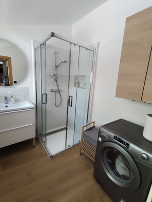Une salle de bain compacte avec une douche Ă parois de verre, un lave-linge et un lavabo blanc avec un miroir.