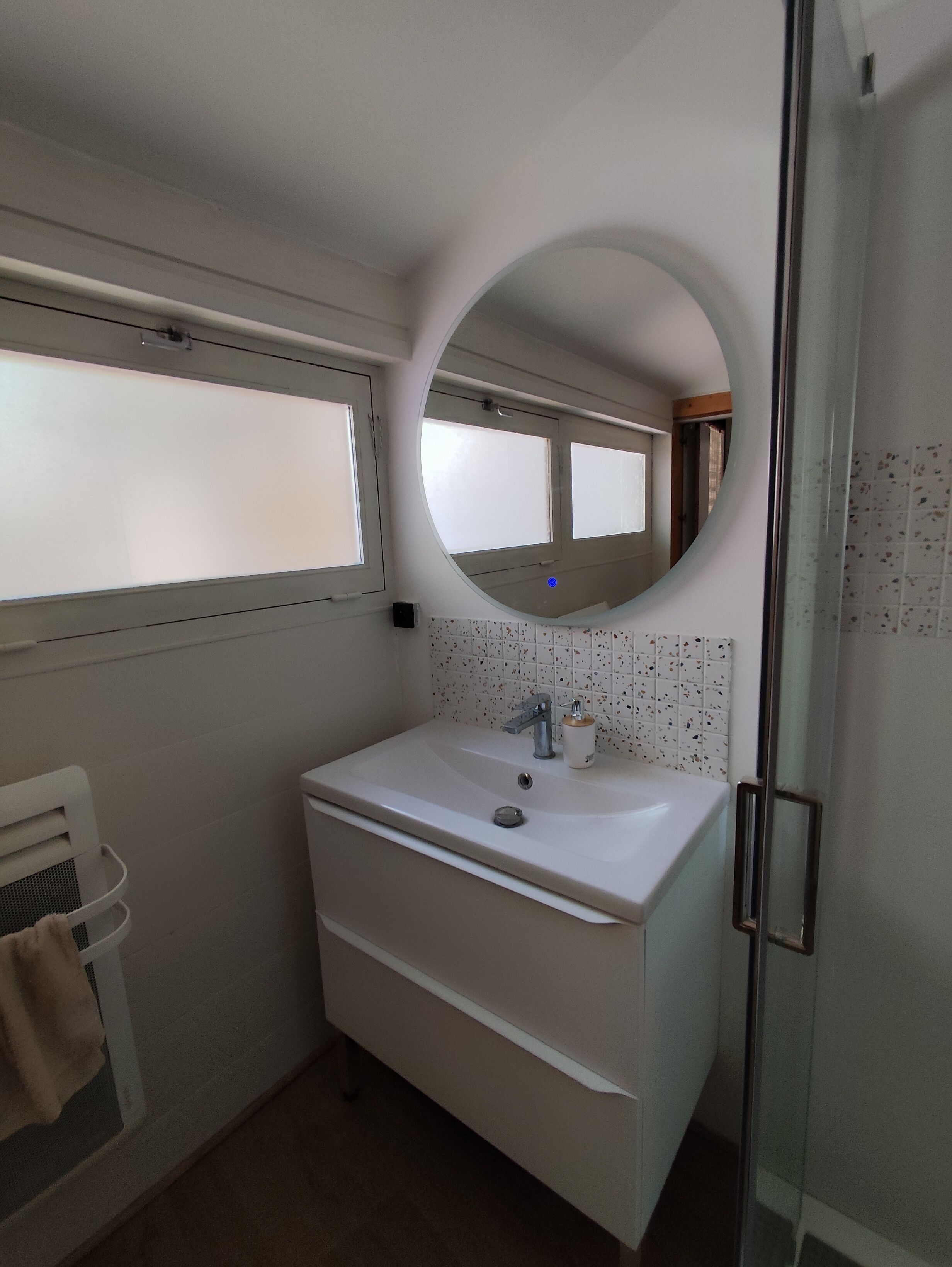 Un baño con un lavabo blanco, un espejo redondo, una ventana y un salpicadero de azulejos.