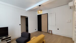 Ein modernes Wohnzimmer mit Fernseher, dunklem Sofa, gelbem Sofa, kleinem Holzschemel und einer Pendelleuchte.