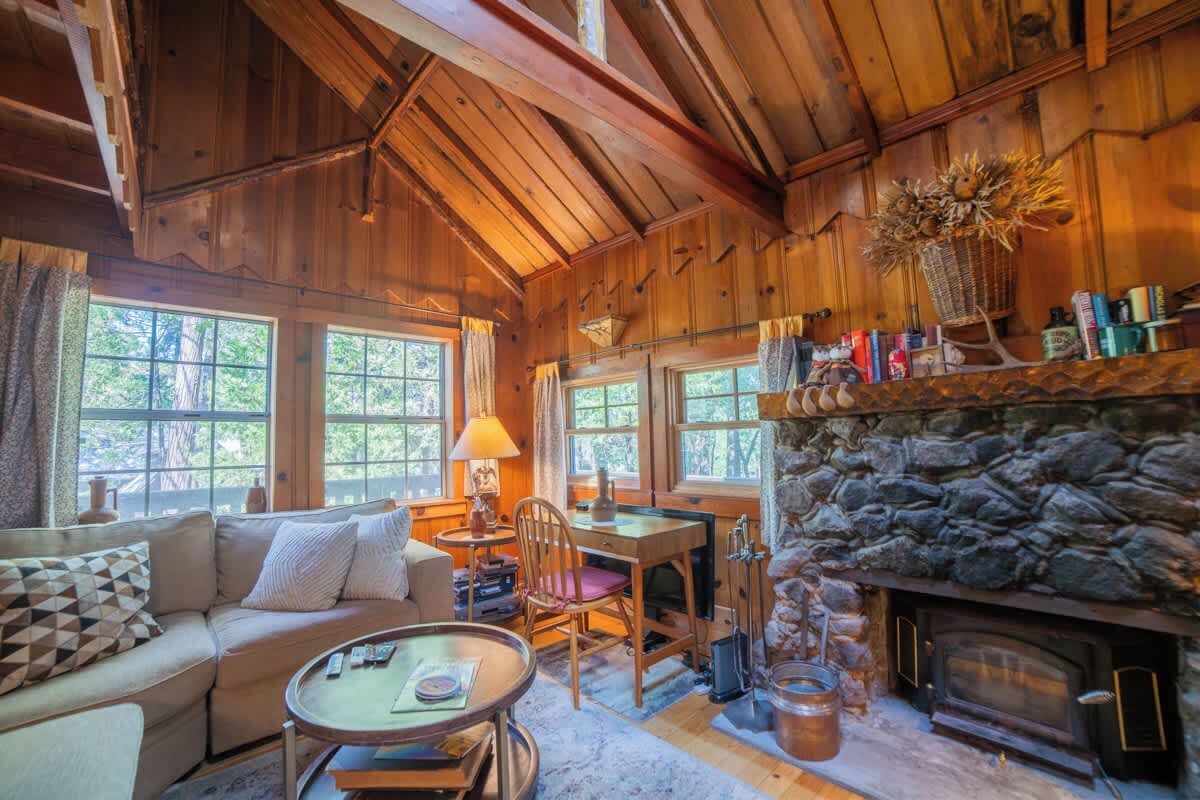 A cozy cabin interior with a stone fireplace, wooden walls, a sofa, a dining table, and a window overlooking greenery.