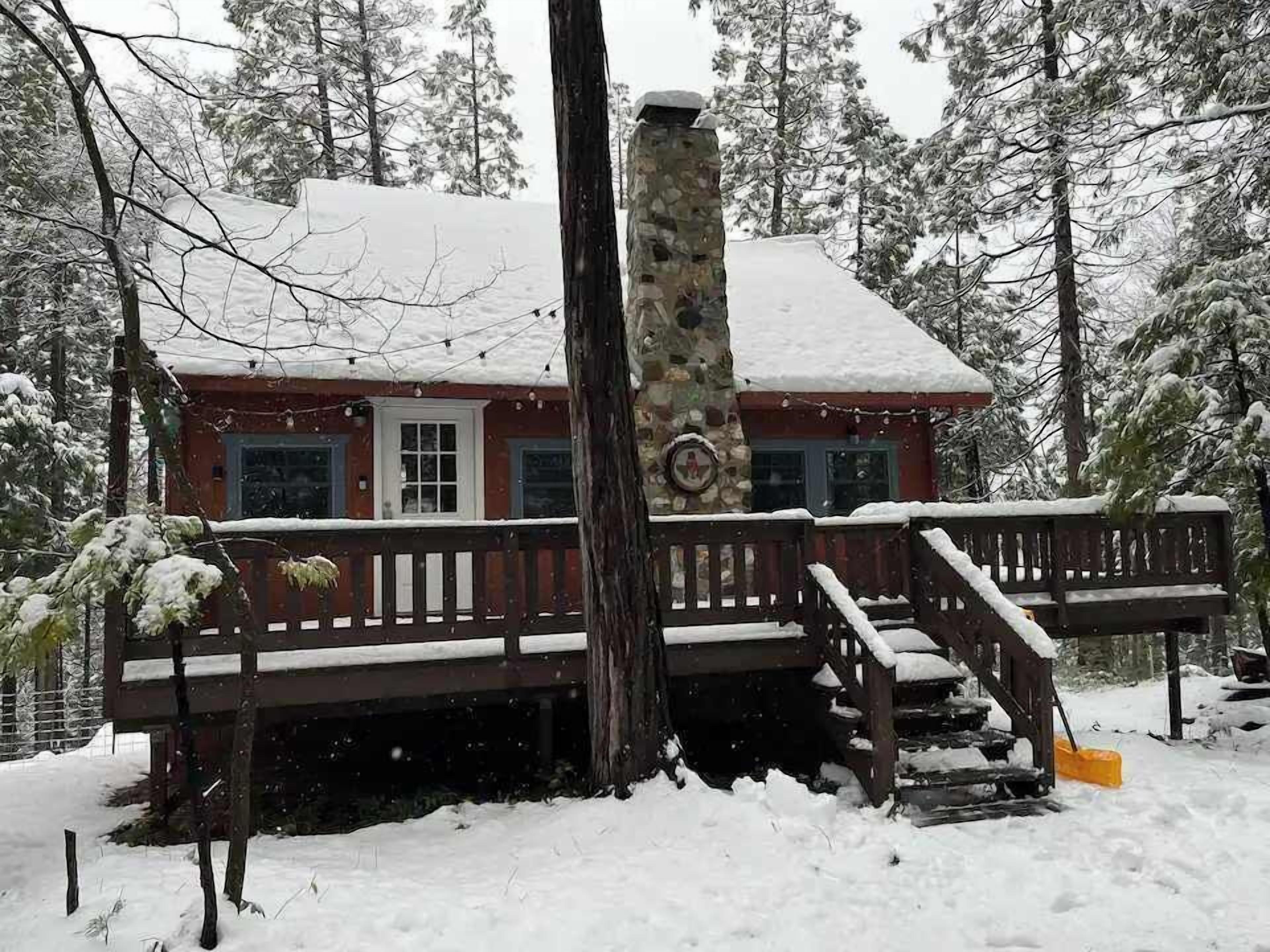 Una cabaña con chimenea de piedra, un porche de madera y un techo cubierto de nieve.