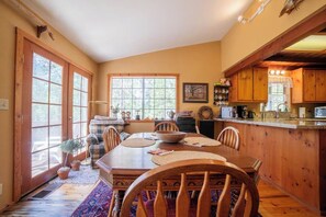 A dining area with a wooden table and chairs, a kitchen with wooden cabinets, and a large window providing natural light. - Daisy’s Place | Cozy Twain Harte Cabin w/ Twain Harte Lake Access (Twain Harte)