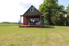 A small, single-story house with a red door and a porch, surrounded by a grassy field and trees.