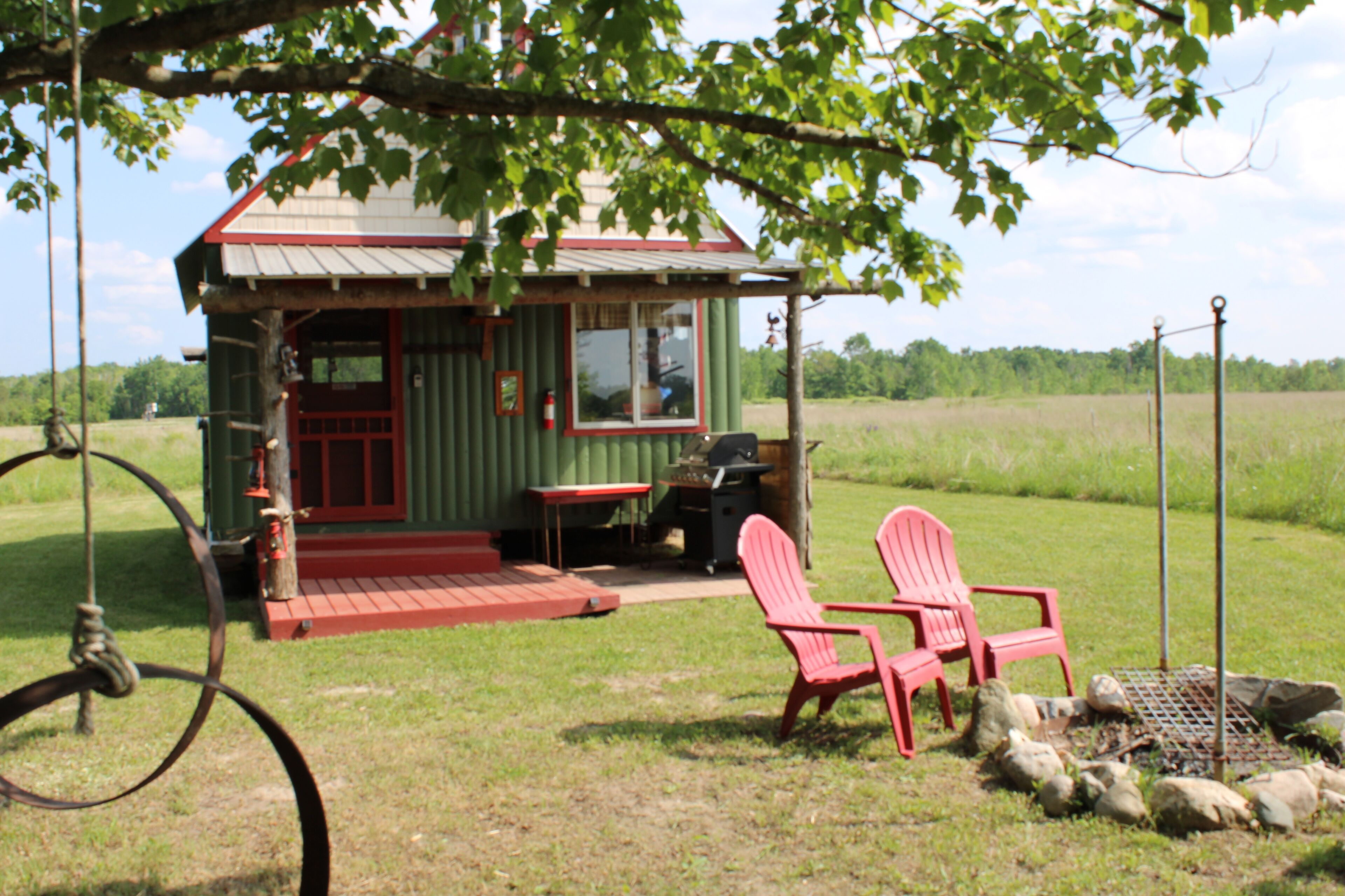 A small green cabin with a porch, two red chairs, a grill, and a swing set.