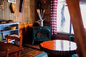 A cozy room with a wooden wall, a green stove, a bookshelf, and a window with a plaid curtain.