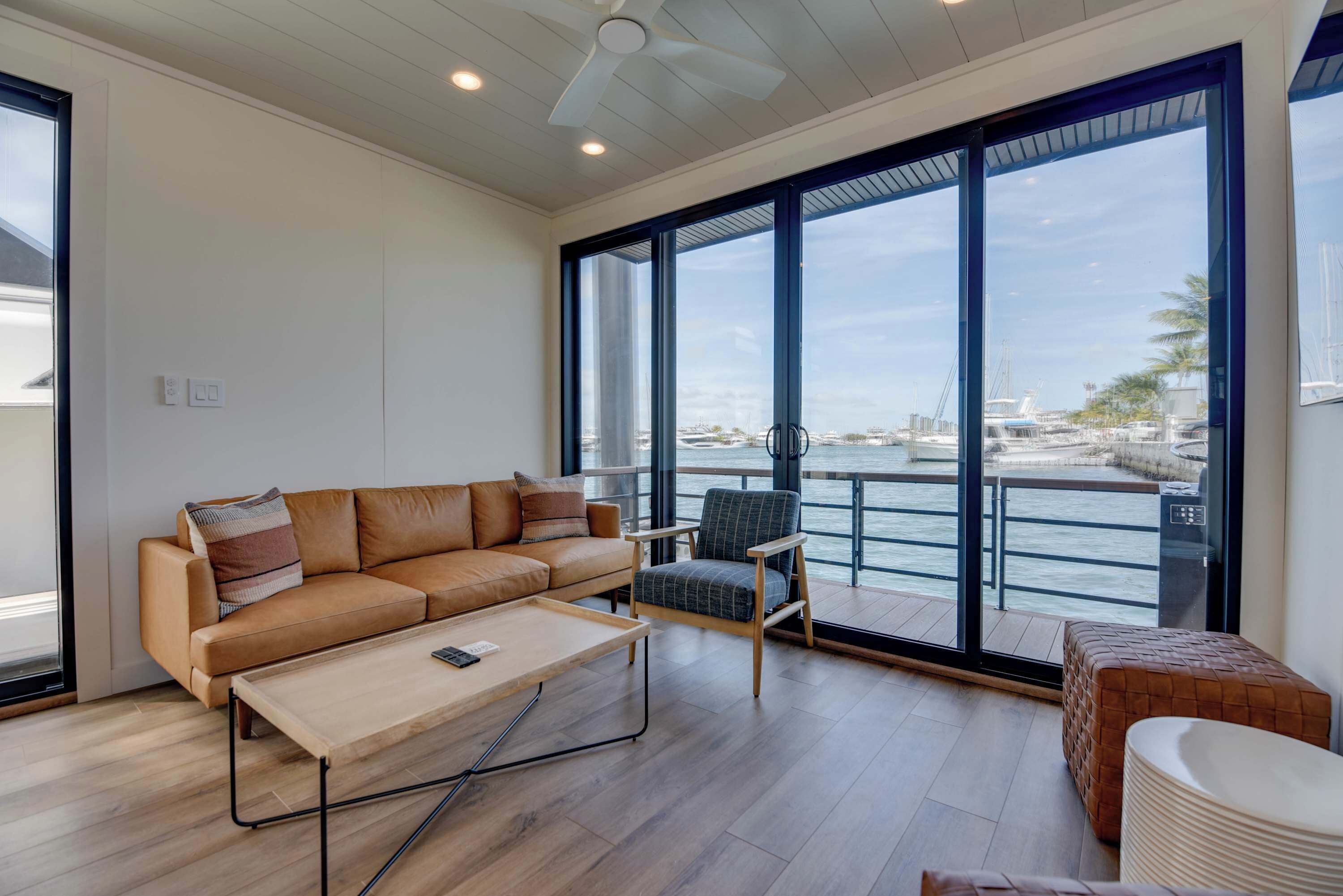 Een moderne woonkamer met een bank, fauteuil en salontafel, grote ramen met uitzicht op de jachthaven, en een plafondventilator.
