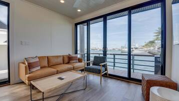 Een moderne woonkamer met een bank, fauteuil en salontafel, grote ramen met uitzicht op de jachthaven, en een plafondventilator.