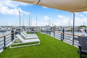 Un puerto deportivo con barcos, una terraza con césped y sillones de descanso, y un cielo despejado.