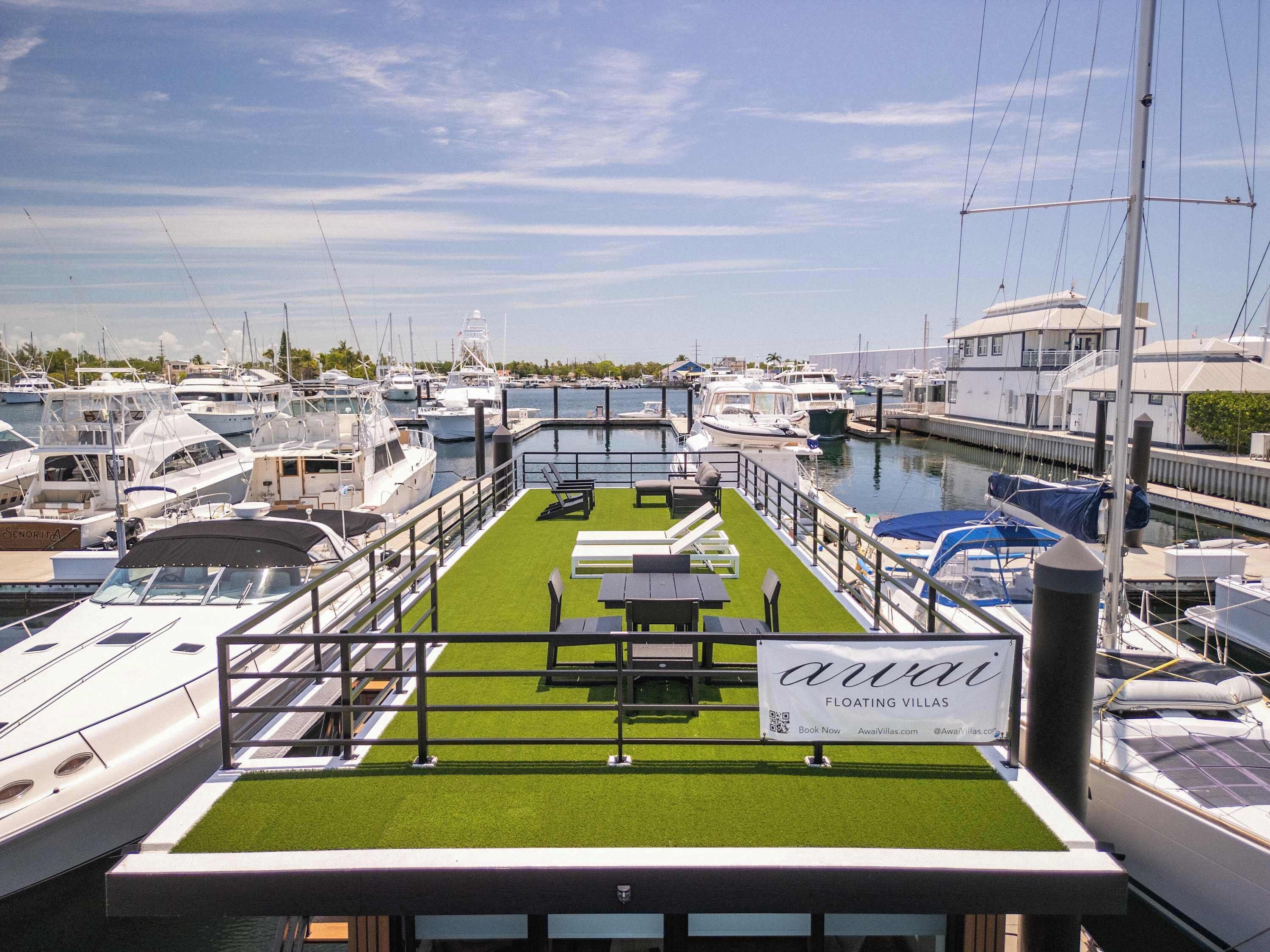 A marina with boats docked, a floating villa with a green deck and lounge chairs, and a sign advertising 'AwaVi' floating villas.