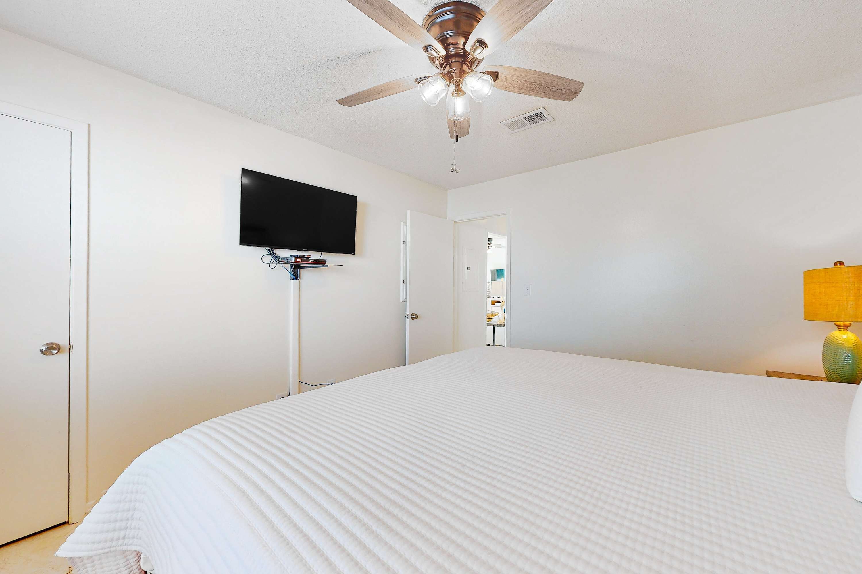 Une chambre à coucher avec un lit, un ventilateur de plafond, un téléviseur fixé au mur et une porte donnant sur une autre pièce.