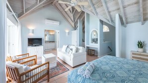 Una habitación amplia con techo alto, cama con ropa de cama estampada, sofá blanco, banco de madera, televisor y zona de cocina.