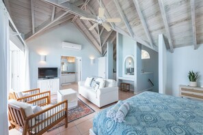 A spacious room with a high ceiling, a bed with patterned bedding, a white sofa, a wooden bench, a television, and a kitchen area.