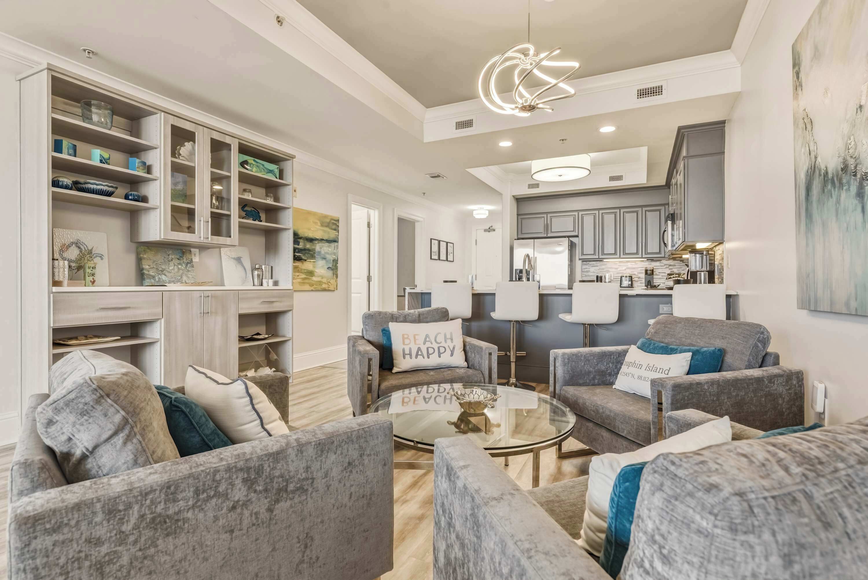 A modern living room with grey upholstered furniture, a glass coffee table, and a kitchen area with grey cabinets and a white countertop.