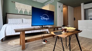 A hotel room with a bed, a TV displaying Disney+, a wooden bench, a table with snacks, and a woven rug. - Sensai Downtown by Riviera Maya Suites  (Playa del Carmen)