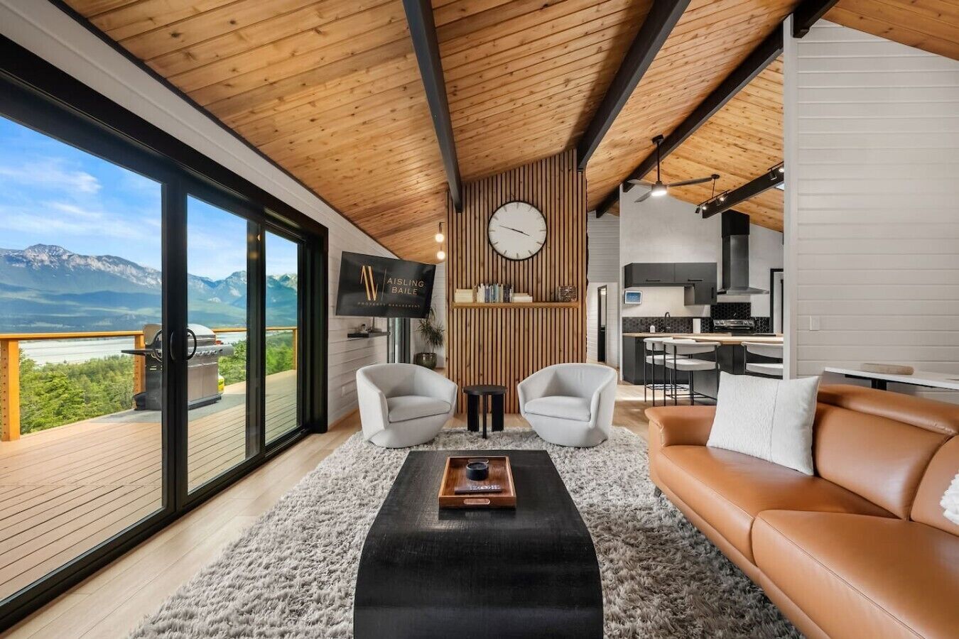 A modern living room with a large window, a wooden ceiling, a sofa, two armchairs, a coffee table, and a kitchen area.