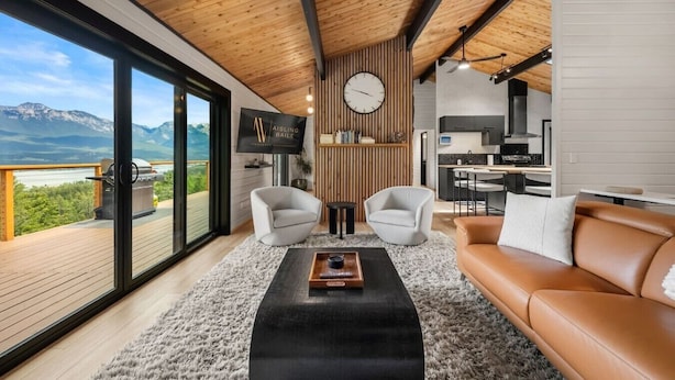 Ein modernes Wohnzimmer mit großem Fenster, Holzboden, einer Couch, zwei Sesseln, einem Couchtisch und einer Küchenzeile.