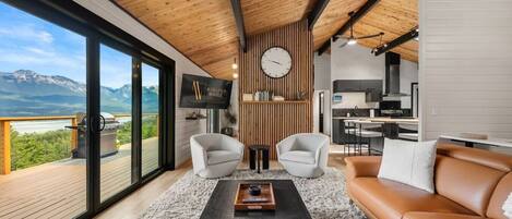 A modern living room with a large window, a wooden ceiling, a sofa, two armchairs, a coffee table, and a kitchen area.