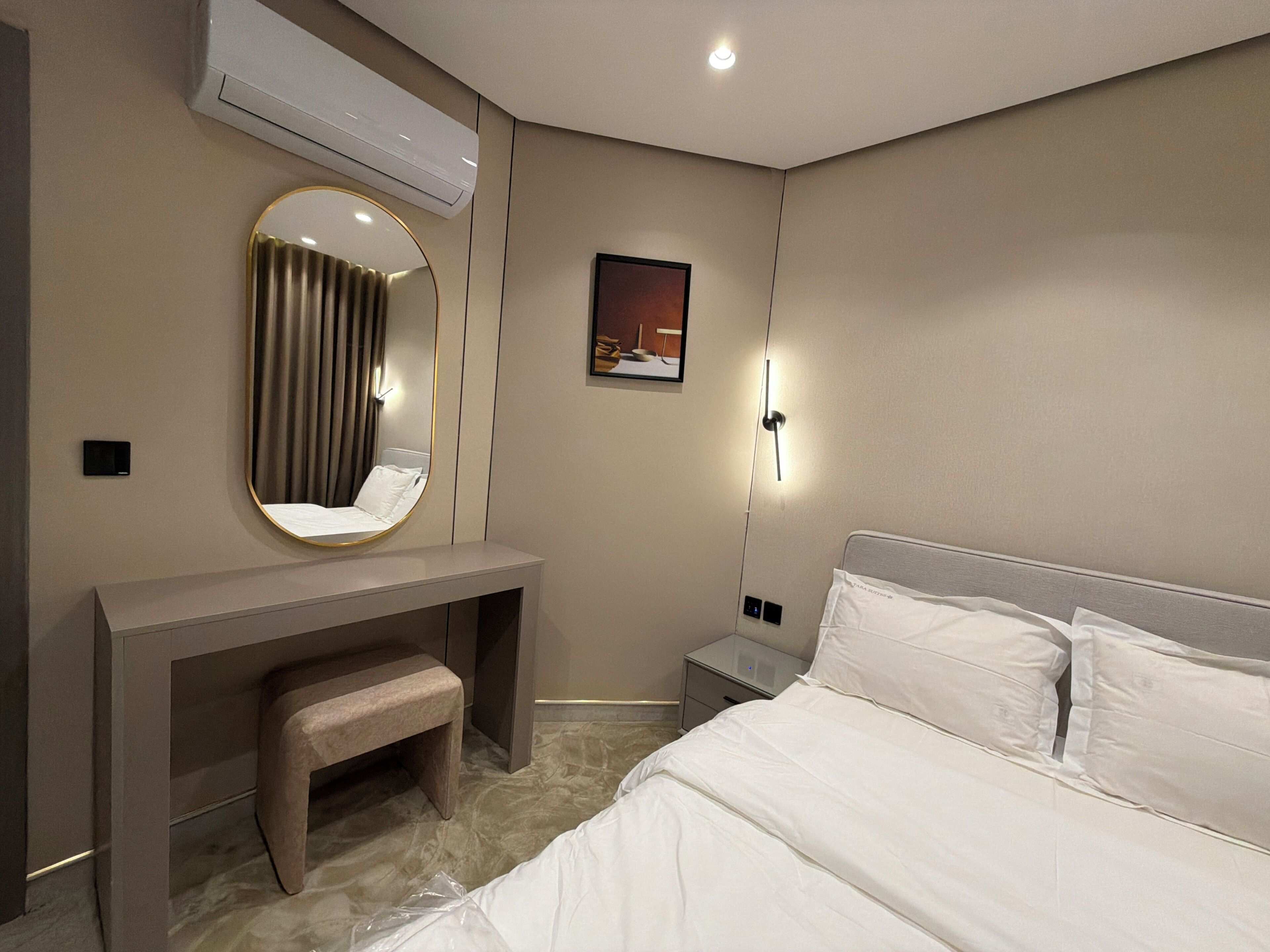 A hotel room with a bed, a bedside table, a desk with a chair, a mirror, an air conditioning unit, a wall-mounted light, and a painting.