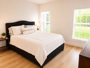 Quarto com cabeceira preta, roupa de cama branca, mesa de cabeceira com abajur, uma pequena planta e vista para um jardim verde pela janela.