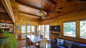 A wooden cabin interior with a dining table, chairs, a sofa, a TV, and large windows.