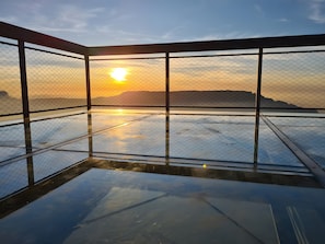 Una habitación con piso de vidrio y vista al atardecer sobre un paisaje montañoso.