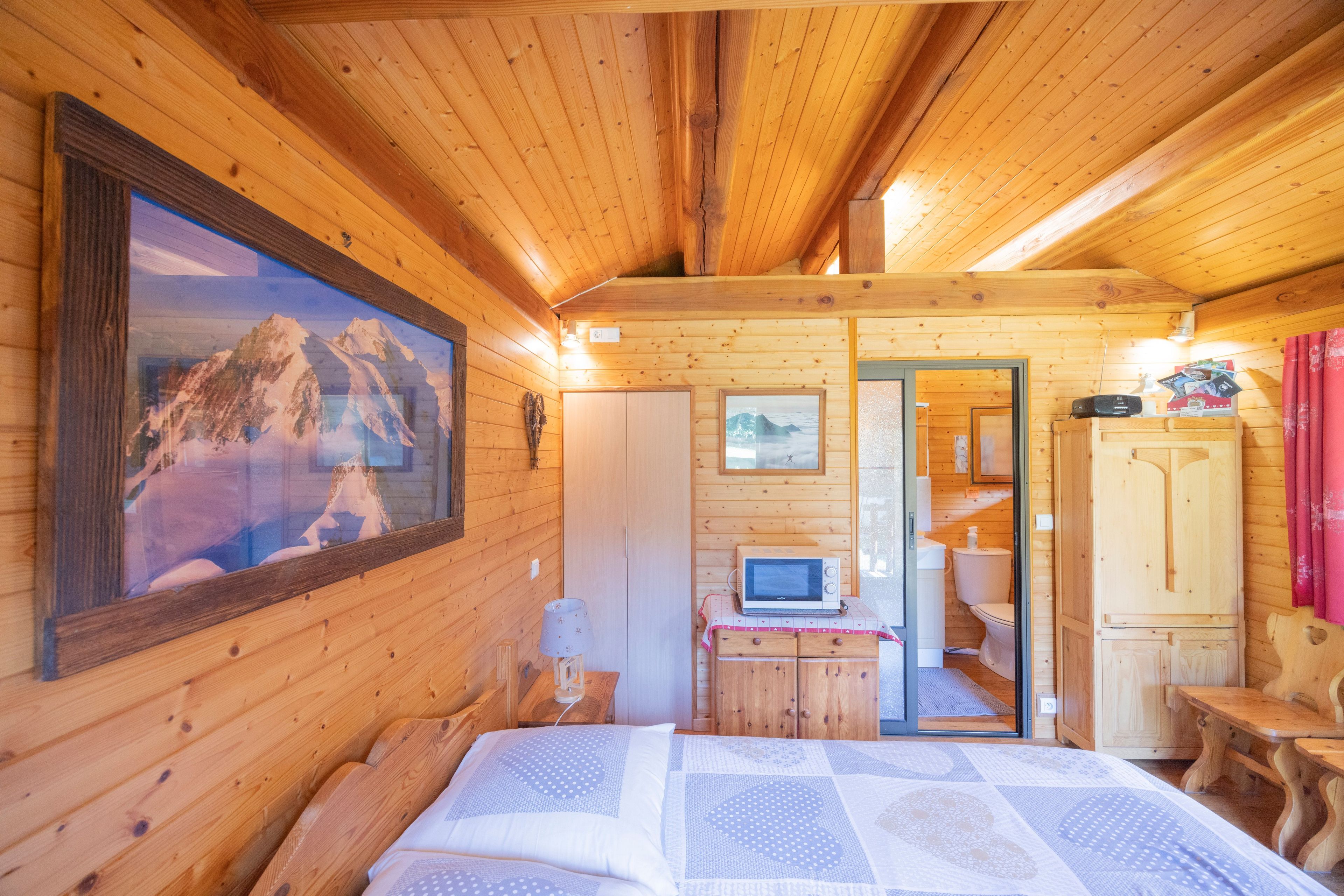 A wooden cabin interior with a bed, a painting of mountains, a small kitchenette, and a bathroom.