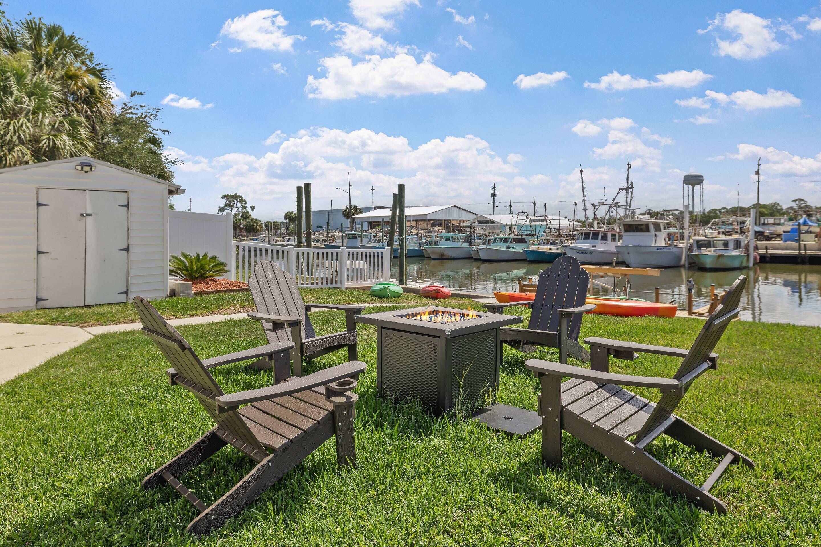 A grassy outdoor area with Adirondack chairs, a fire pit, and a marina with boats in the background.